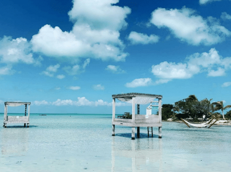 Overwater Cabanas in Belize Overwater cabanas on stilts in shallow turquoise waters, with a small island, palm trees, and hammocks in the background under a bright blue sky with fluffy clouds.