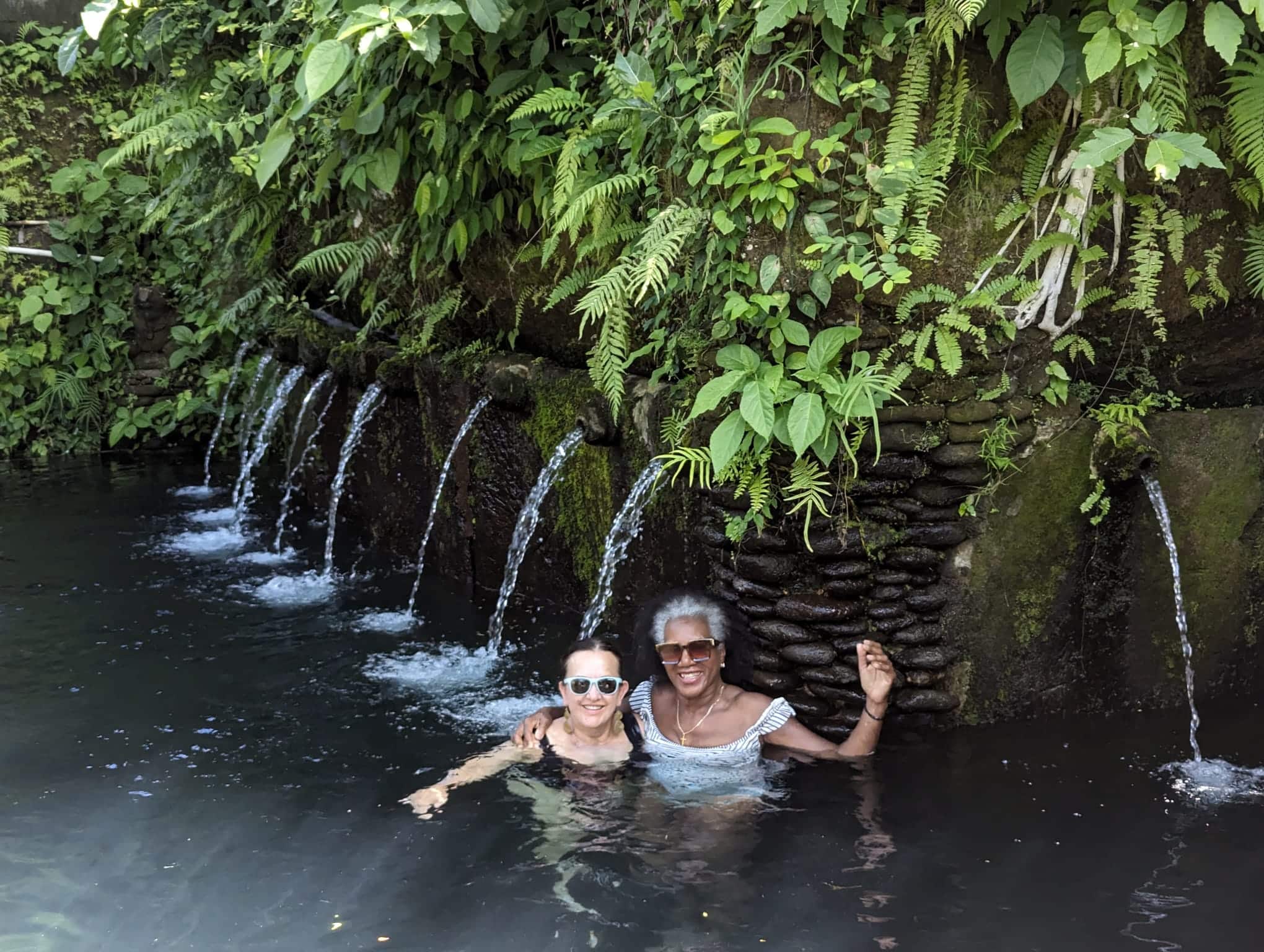 WTA travelers smiling and soaking in a hot spring in Bali, surrounded by small stone fountains and lush tropical plants. WTA travelers smiling and soaking in a hot spring in Bali, surrounded by small stone fountains and lush tropical plants.