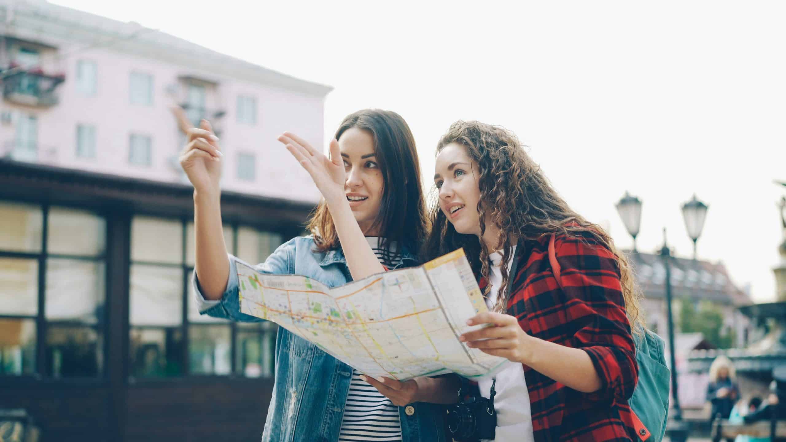 Cheerful young women best friends with backpacks are looking at map and talking standing together in beautiful town in the street and discussing right direction Cheerful young women best friends with backpacks are looking at map and talking standing together in beautiful town in the street and discussing right direction